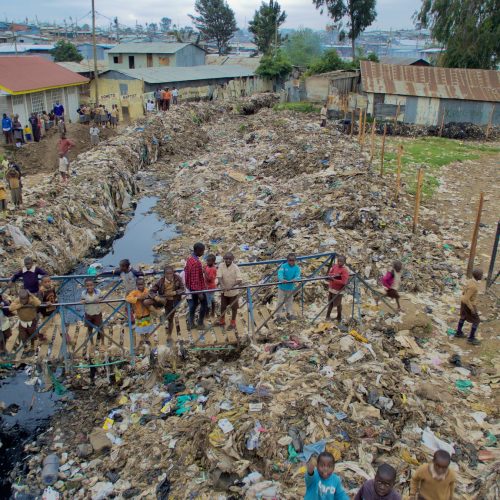 People gathered on a waste-strewn riverbank beside housing
