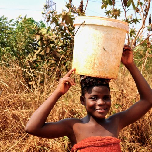 A girl carrying a bucket of water on her head through dry grassland