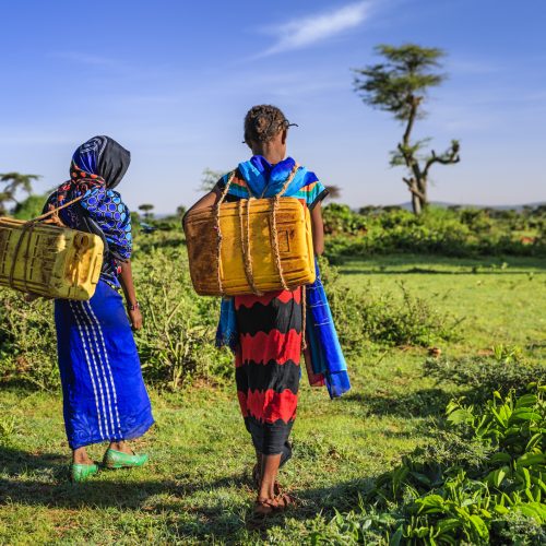 Two women carrying large water containers on their backs across grassland